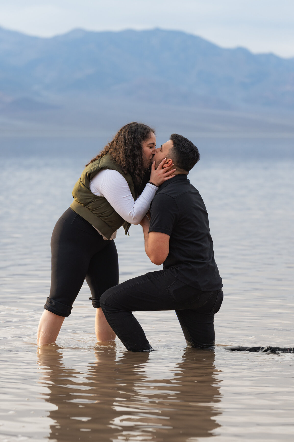 A person kneeling down in shallow water proposing as they kiss their partner
