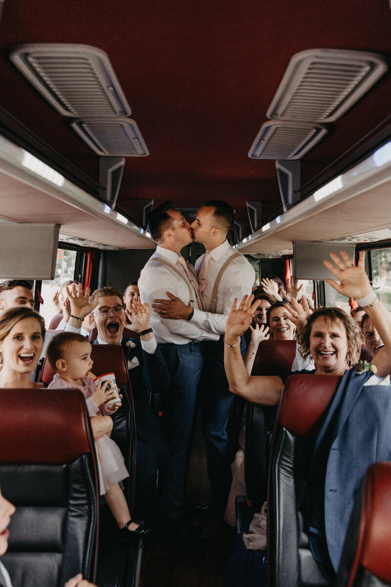 candid moment of grooms kissing with everyone celebrating on their party bus