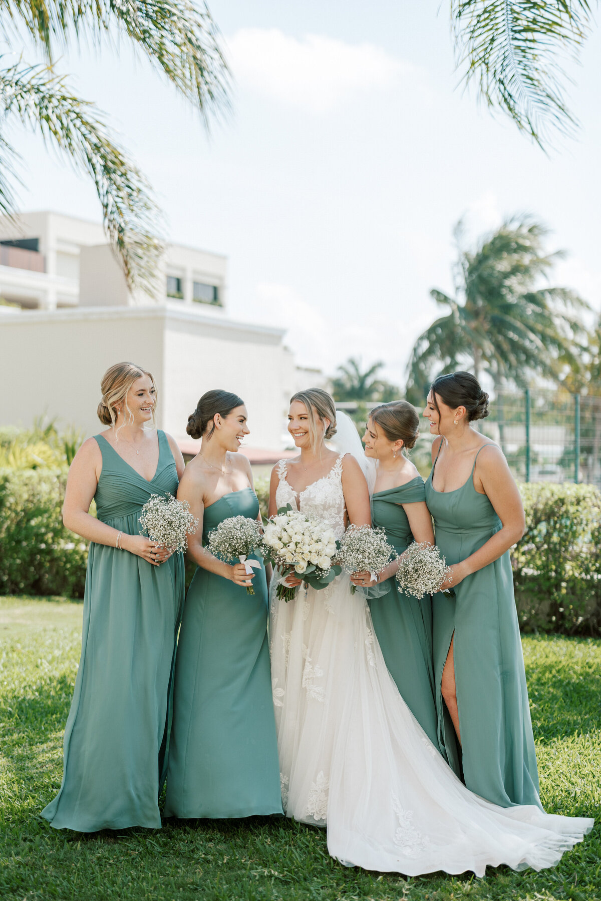 Bride with bridesmaids in sage green dresses holding white bouquets during Cancun wedding.