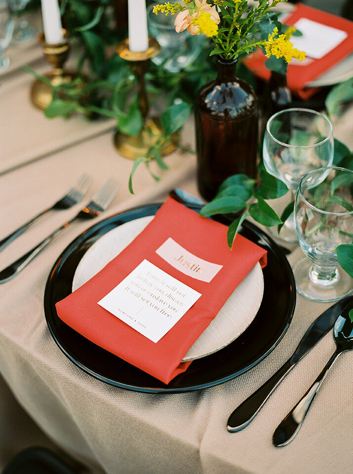 White place card with a gold font set atop a red napkin and white plate with black charger.
