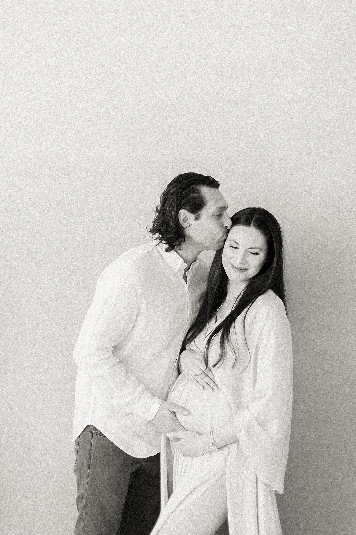 Black and white in studio maternity portrait of an expecting Dallas  mother being held and kissed by her spouse.