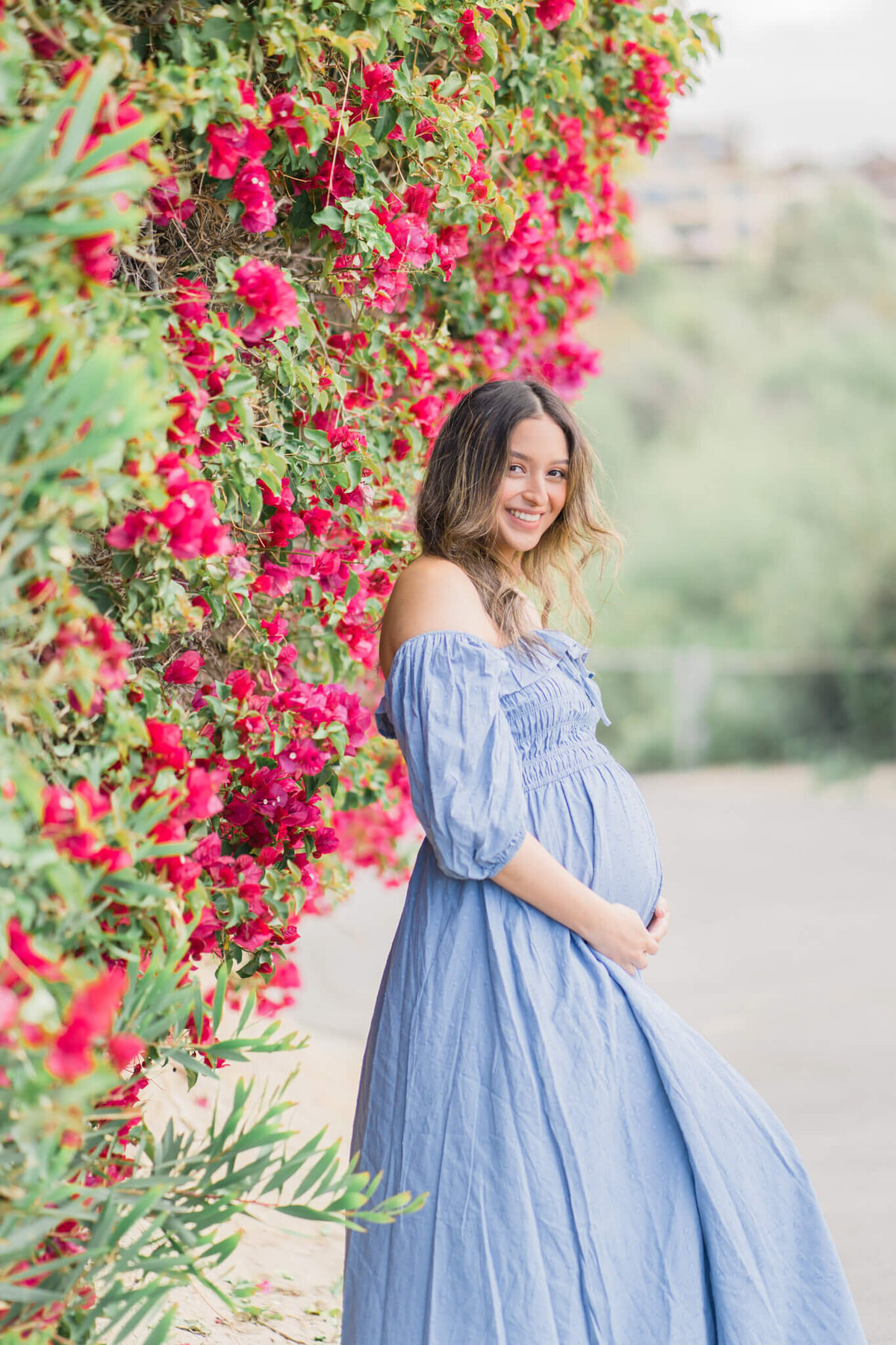 Pregnant mom in blue dress leaning against a wall of pink flowers