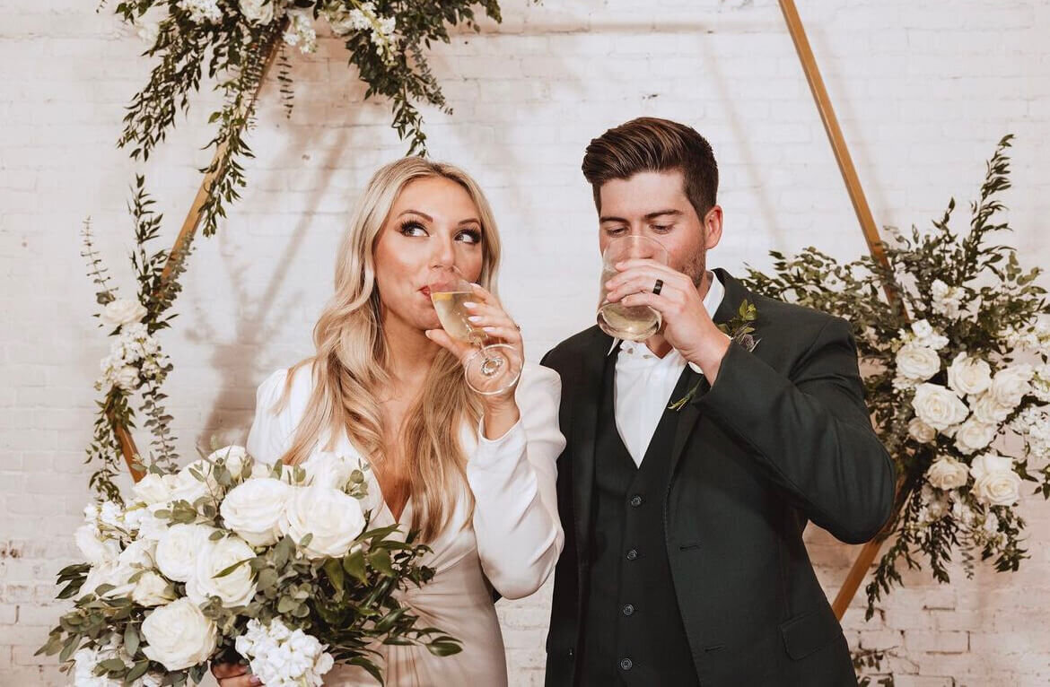 Bride with long curled hair and airbrush makeup holding bouquet, groom with clean-cut style enjoying a toast | San Antonio wedding hair and makeup artist