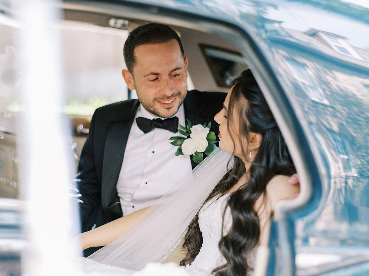 A bride and groom sit inside a car, smiling and looking at each other, capturing the joy of their classic Calgary wedding.