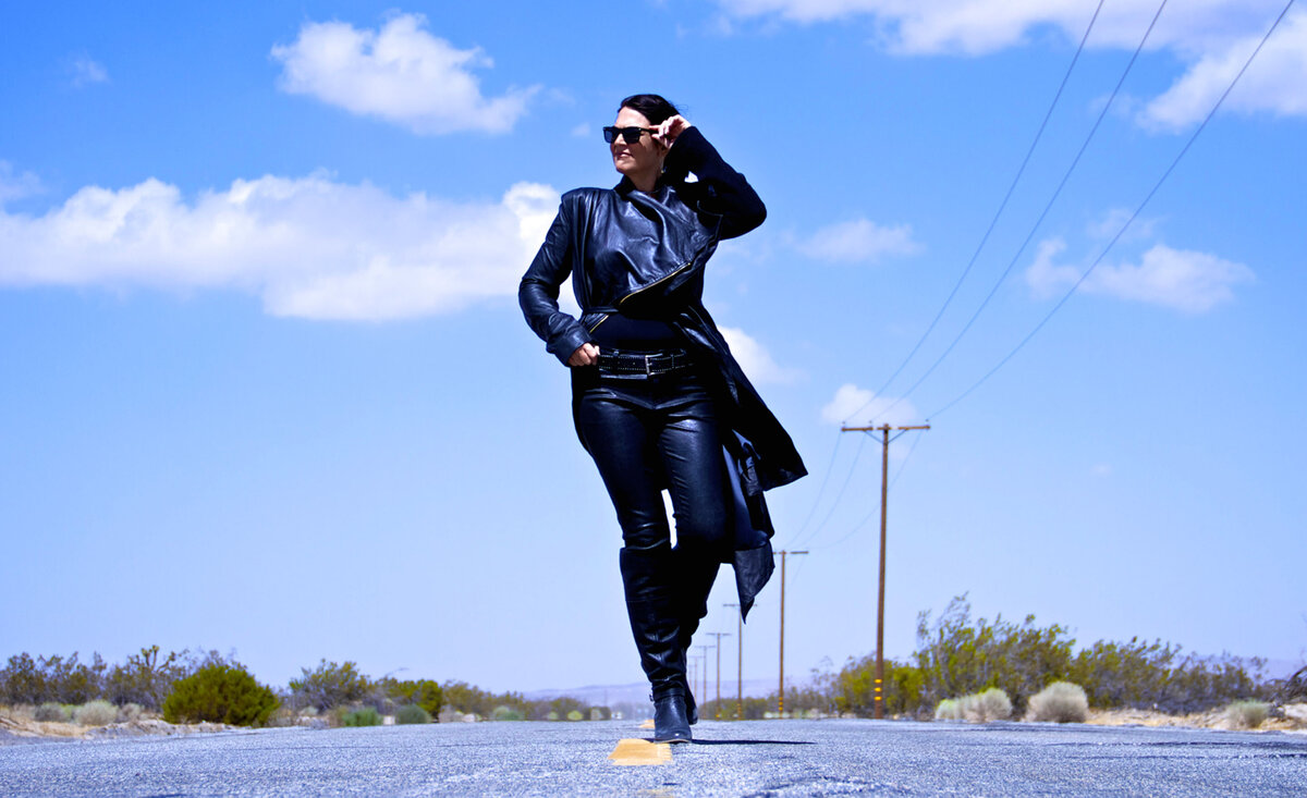 Female musician portrait Leslie Cours Mather walking down desert highway wearing black leather outfit with sunglasses