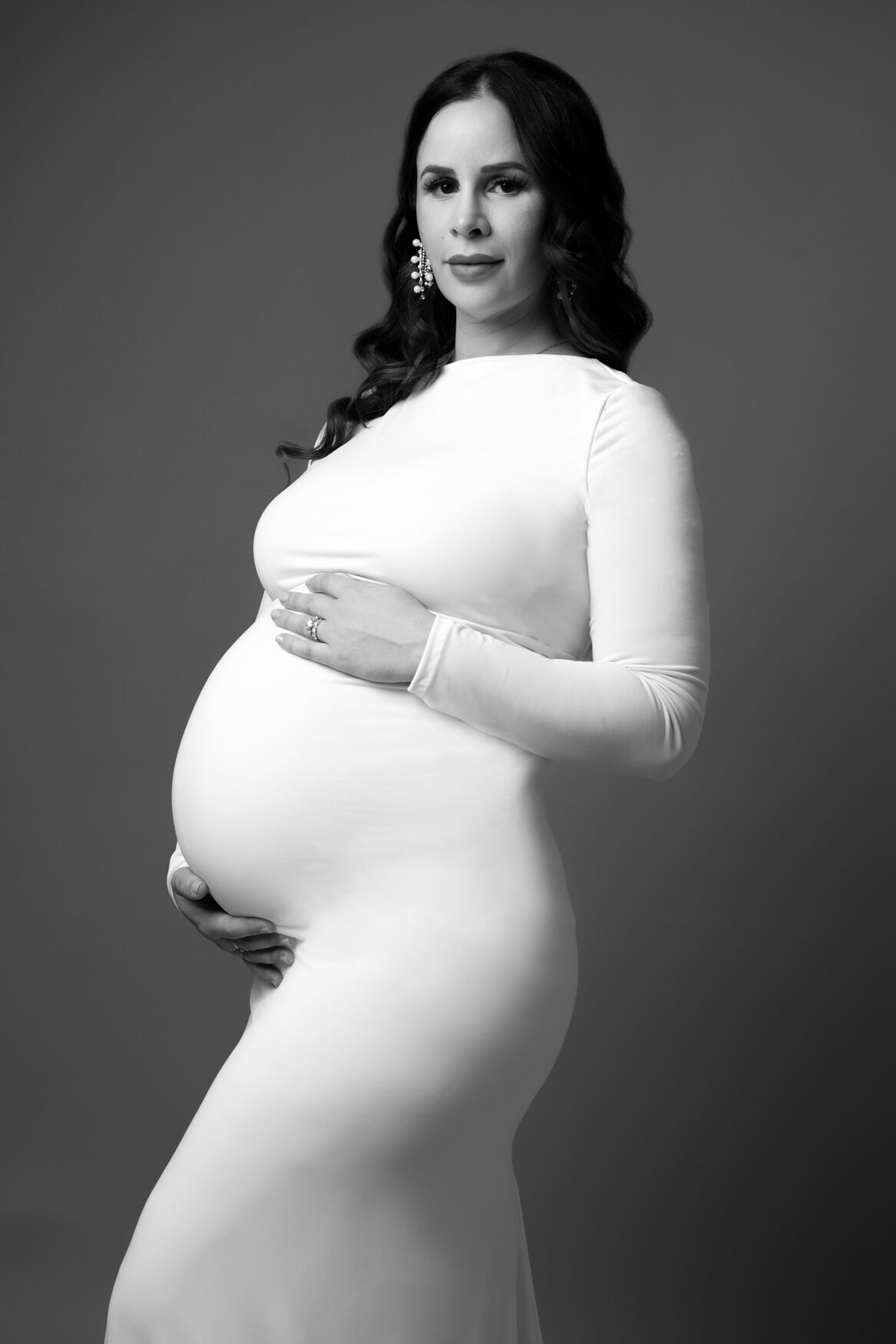 A maternity portrait of a woman wearing a white long sleeve thigh dress looking to the camera and touching her belly with both hands