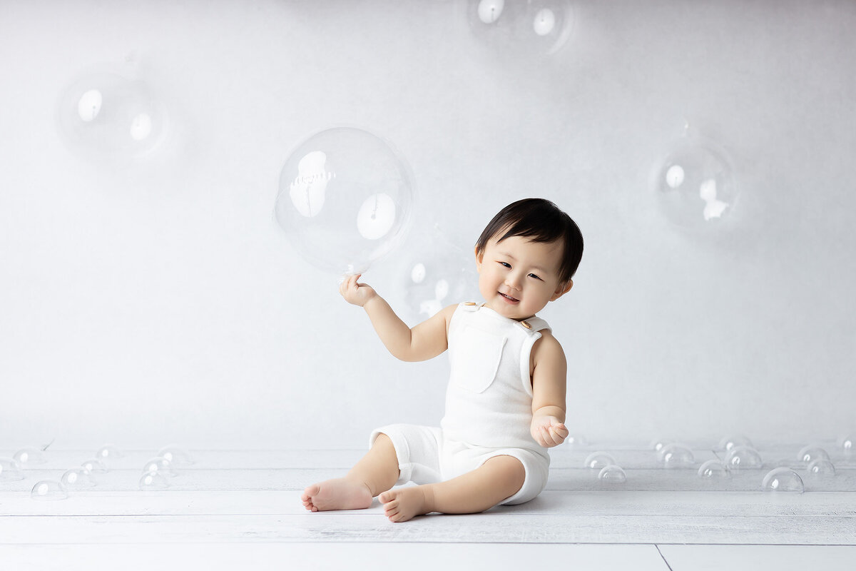 Dallas milestone photographer captures a joyful baby wearing a white romper, sitting on a clean backdrop surrounded by bubbles, with a big smile as they play with a large bubble in the air.