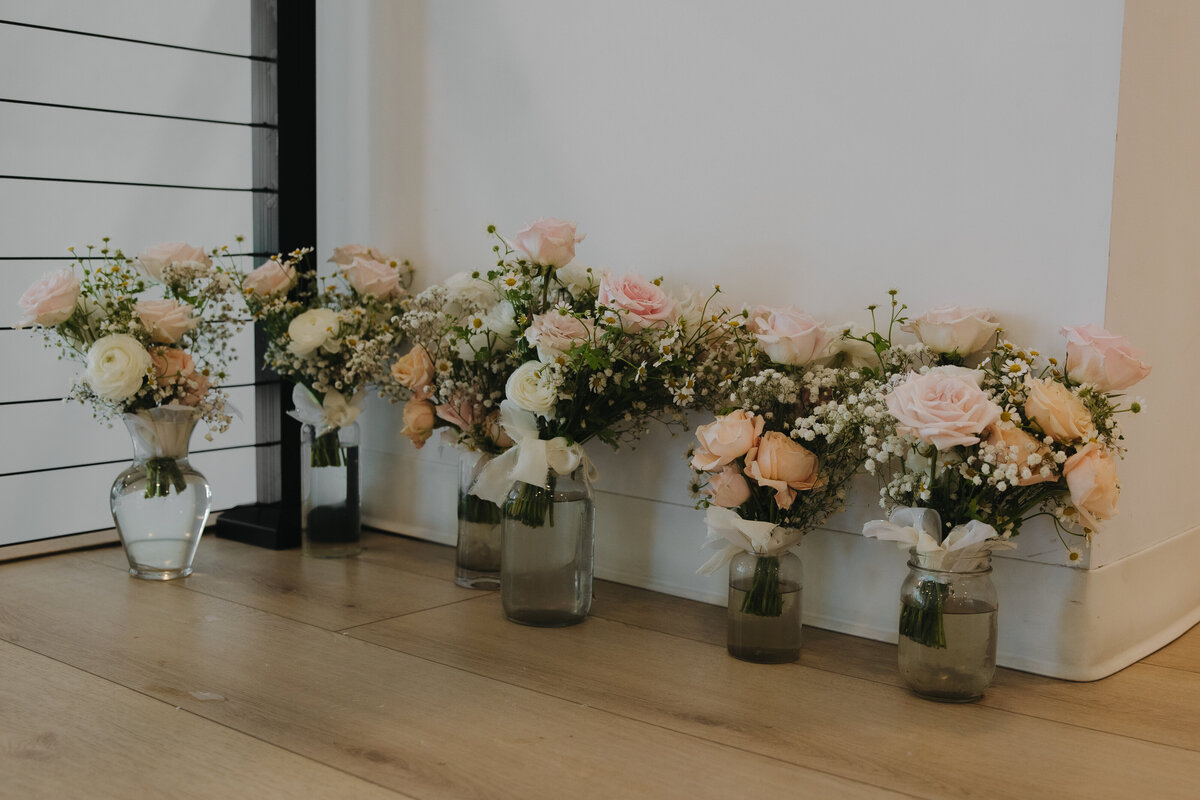 Bridesmaid bouquets in clear vases with water. There are white linen bows tied around each bouquet. The flowers are light pink and light orange roses with white peonies surrounded by baby's breath. The vases are all on a wooden floor up against a white wall and black railing