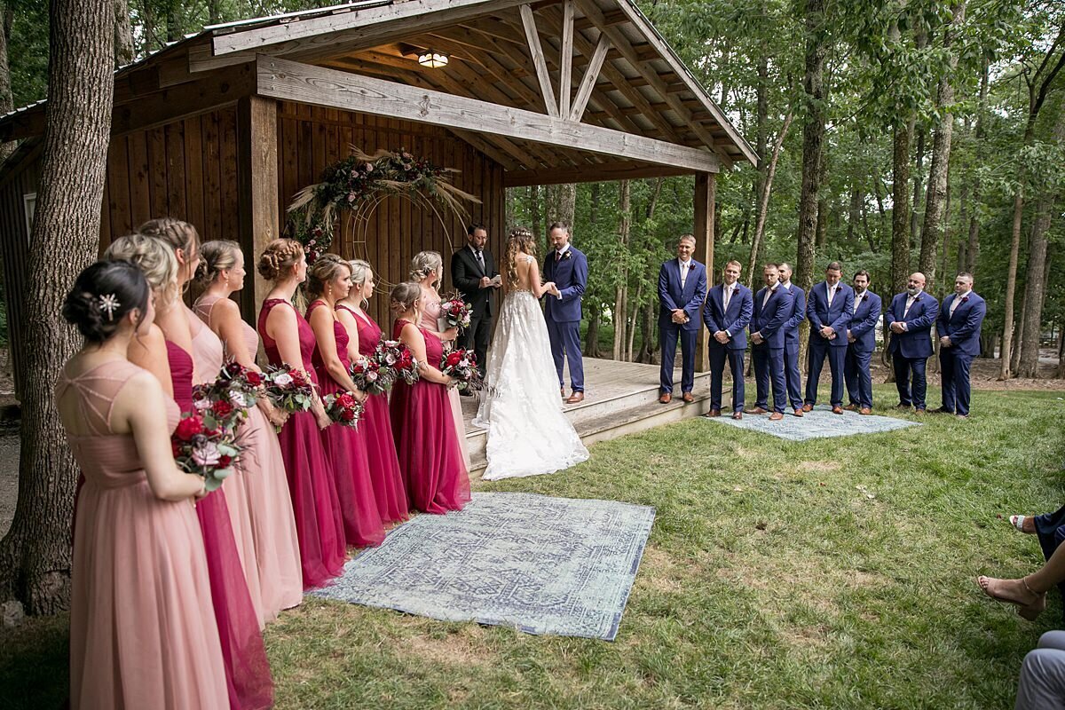 Wedding ceremony at Saddle Woods Farm with groom and groomsmen wearing navy blue suits with burgundy ties and bridesmaids wearing burgundy and blush bridesmaid dresses as they watch the bride and groom get married under the rustic wooden pavilion.