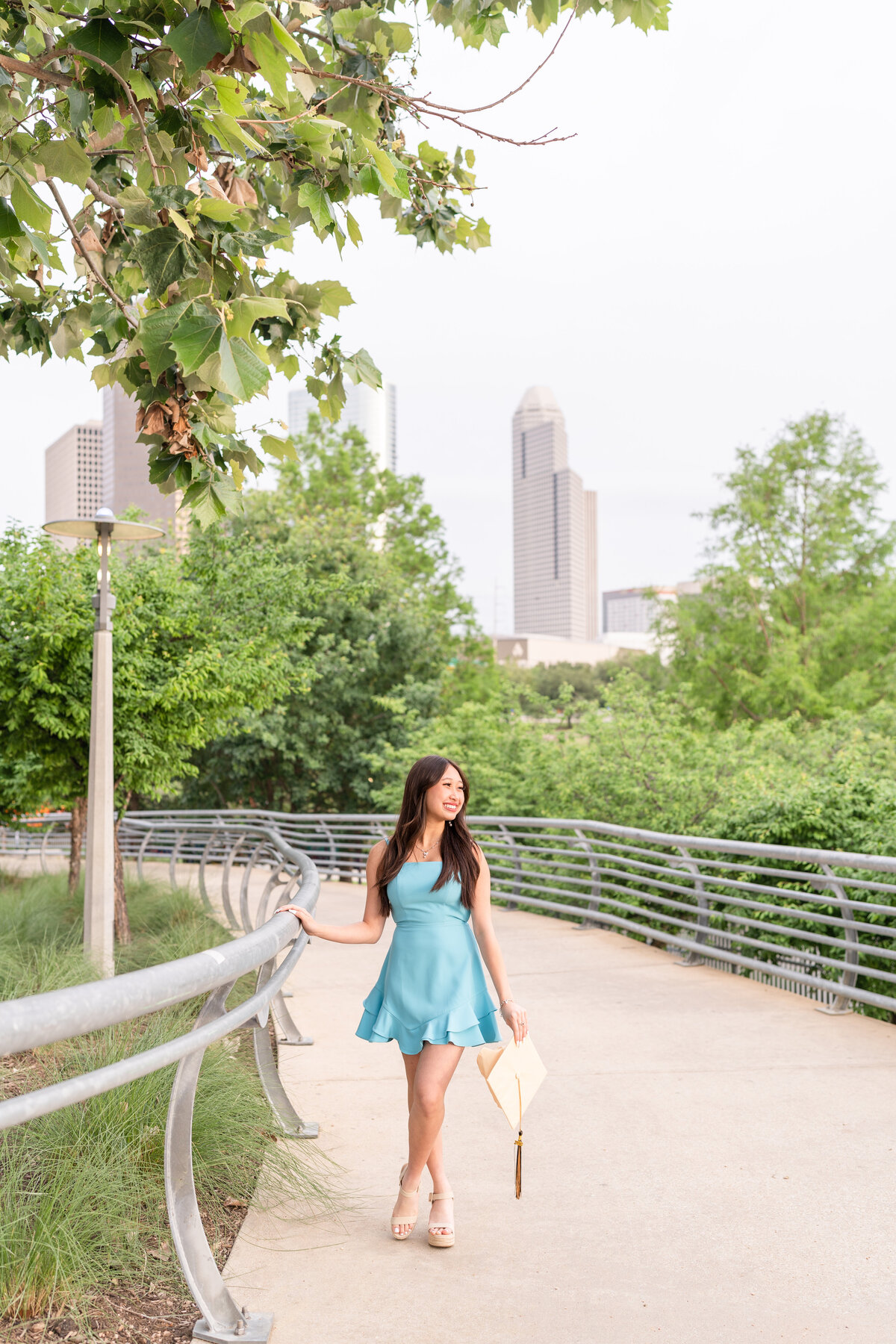 Houston senior girl holding onto railing and holding cap while smiling away in middle of park in Downtown Houston while wearing baby blue dress