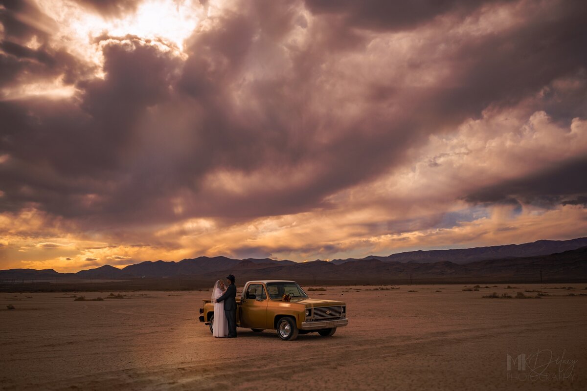 Purple skies with fluffy clouds golden hour sunset elopement photography  dry lake bed mountains Dry lake bed elopement bride in lace wedding gown with glasses groom in black suit white dress shirt with  fedora  classic chevy truck gold  restored with golden hour sunlight las vegas wedding photography las vegas elopement las vegas wedding photographers mk delacy photography