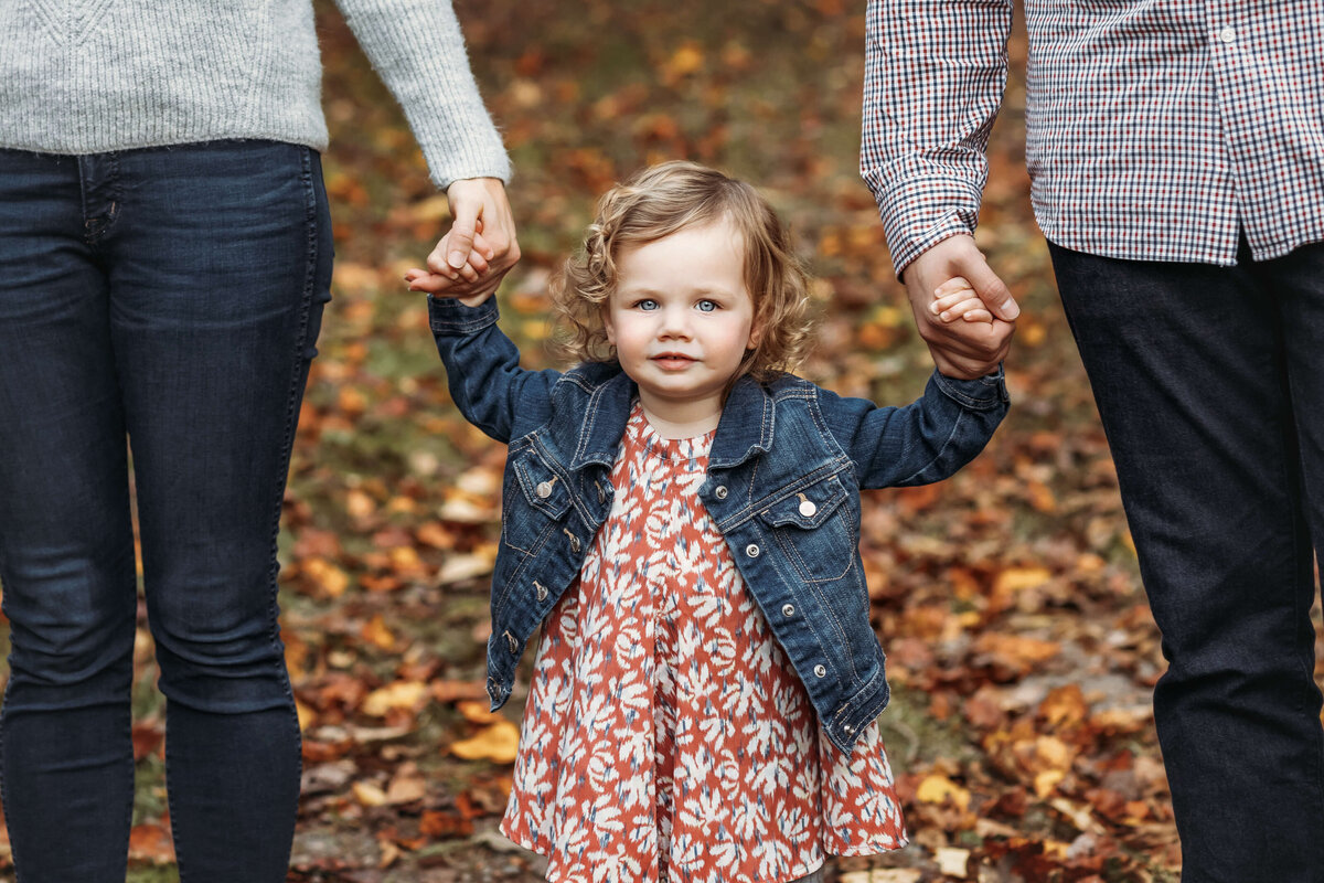 little girl holding mommy and daddy's hands in the fall with pretty leaves behind her