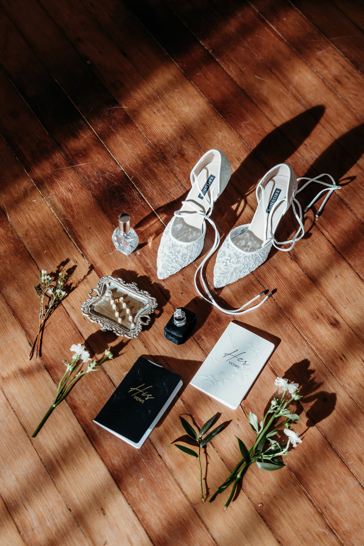 a luxurious details photo of a brides shoes, vow books, rings and perfume in front of a window shadow