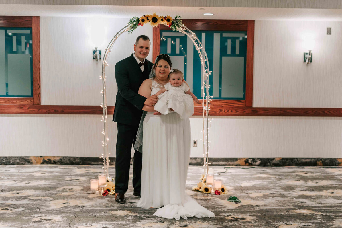 Bride and groom standing  in front of their wedding arch with their daughter.