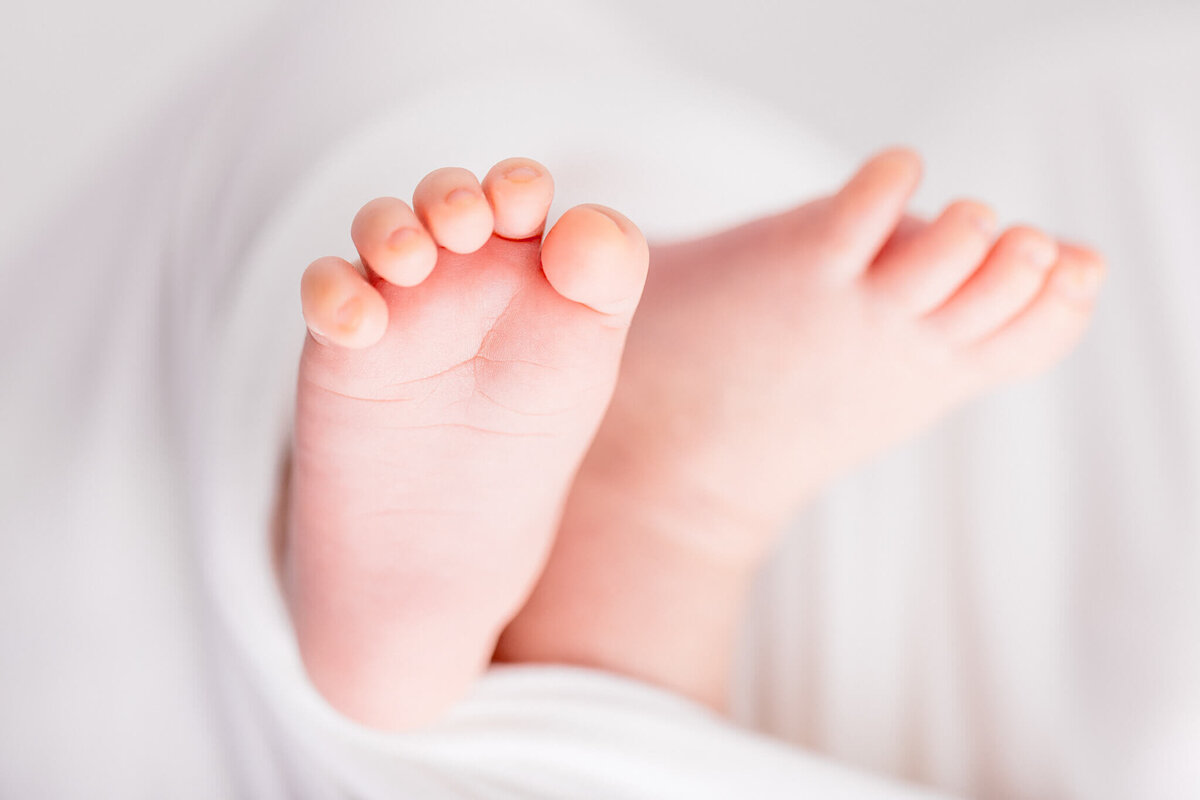 Close-up of a babys feet wrapped in a soft white blanket. The tiny toes are in focus, conveying a sense of warmth and tenderness. The background is blurred, emphasizing the small, delicate details of the babys feet.