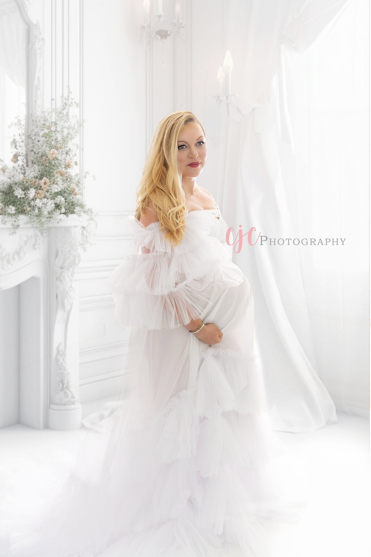 A pregnant blonde woman stands in a studio in a sheer maternity gown hoding her bump