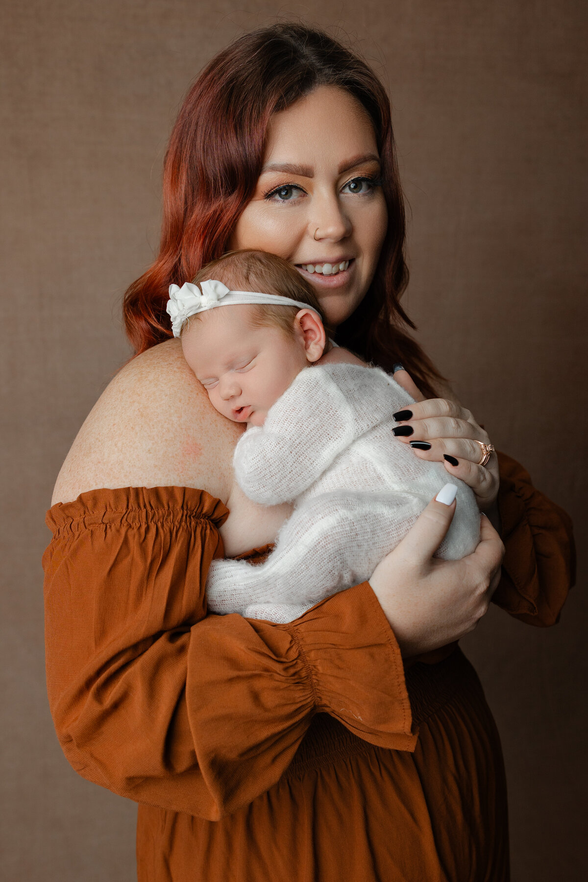 Mother holding her newborn baby girl close, dressed in a soft white knit outfit with a bow headband during their studio session. Captured by Inland Empire newborn photographer Kaitlyn Dawn Photography.