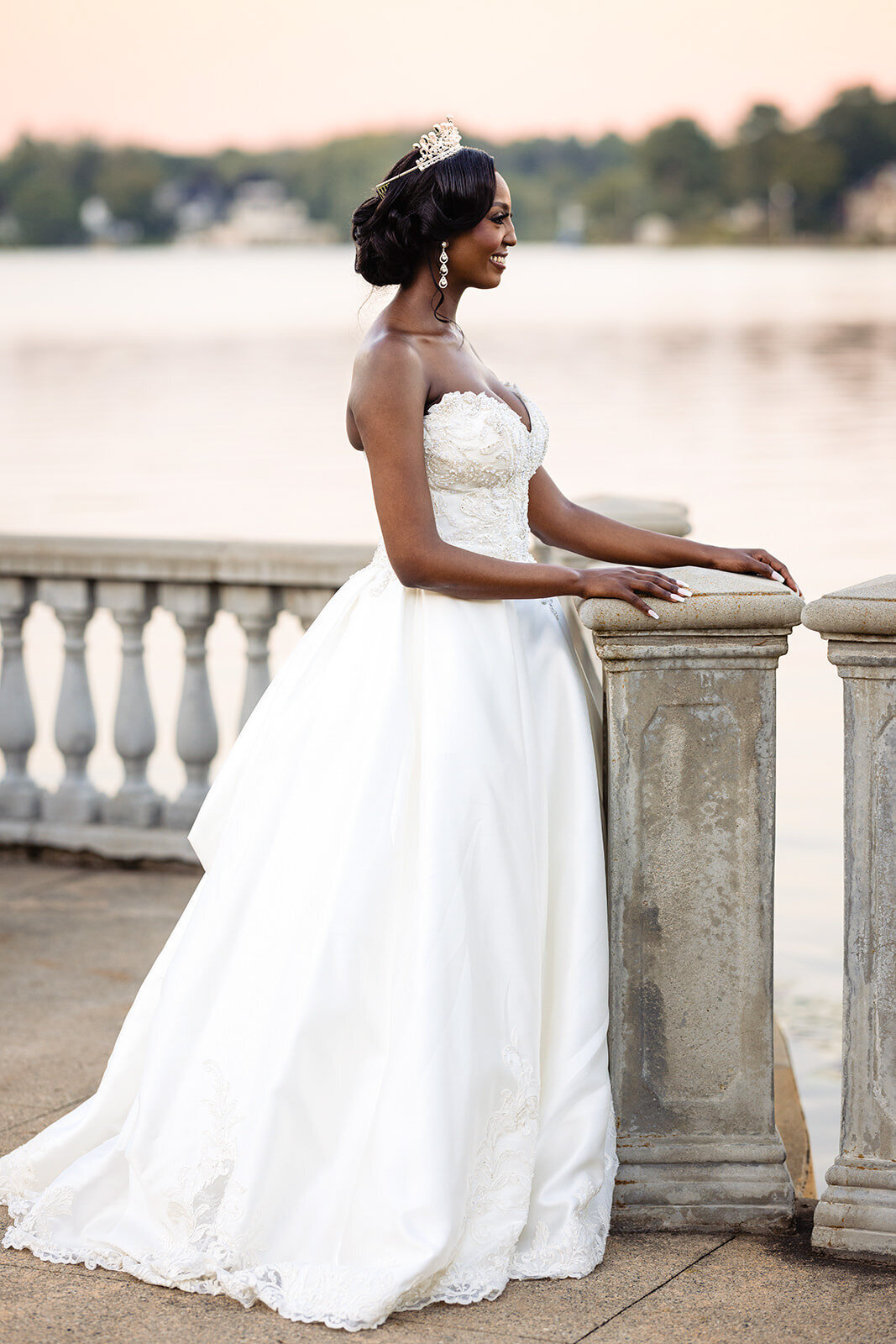 Bride in a strapless wedding gown with a tiara, gazing out over a lake at sunset