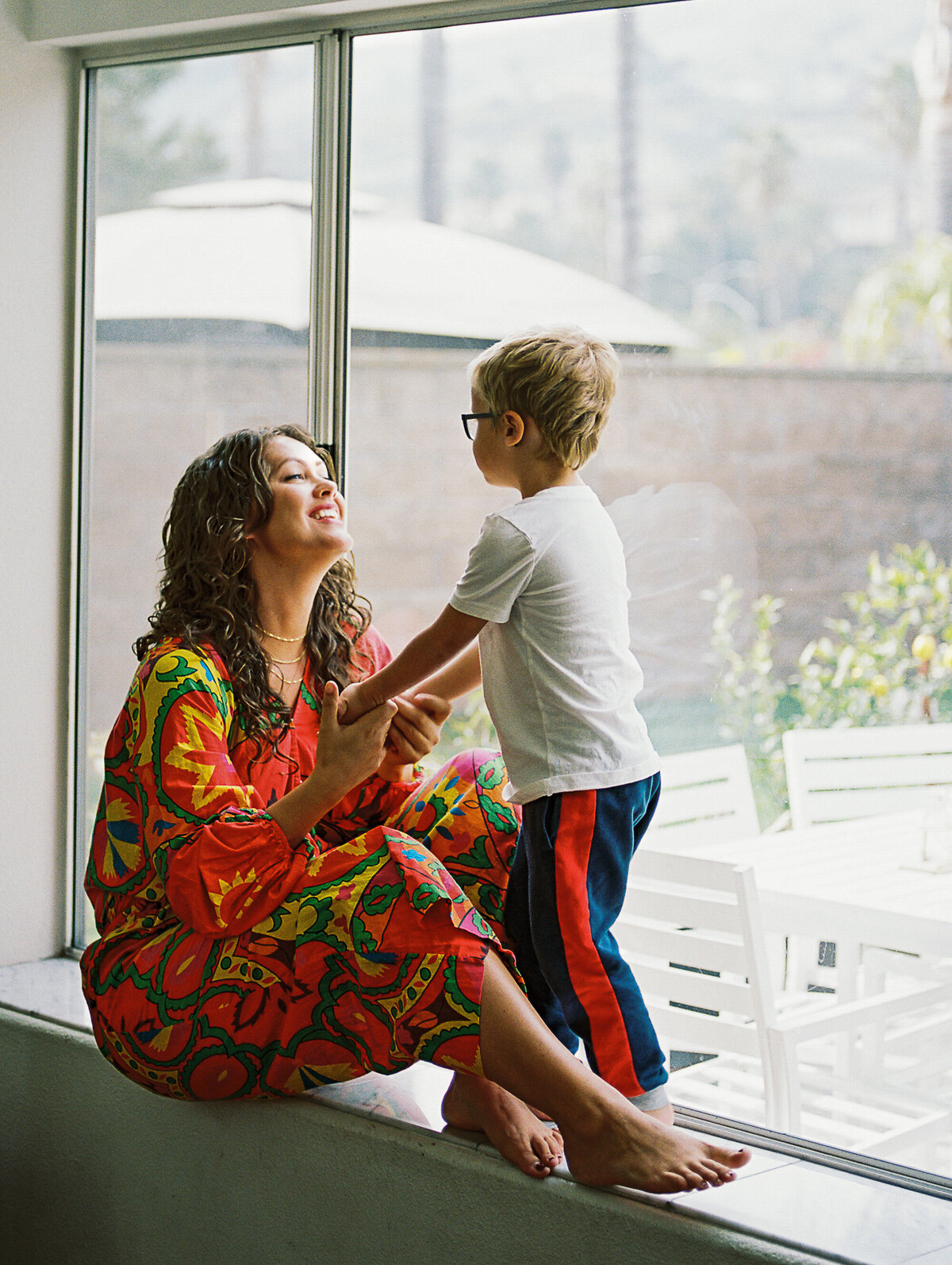 a mom in a red jumpsuit sits on a window ledge and looks at her son