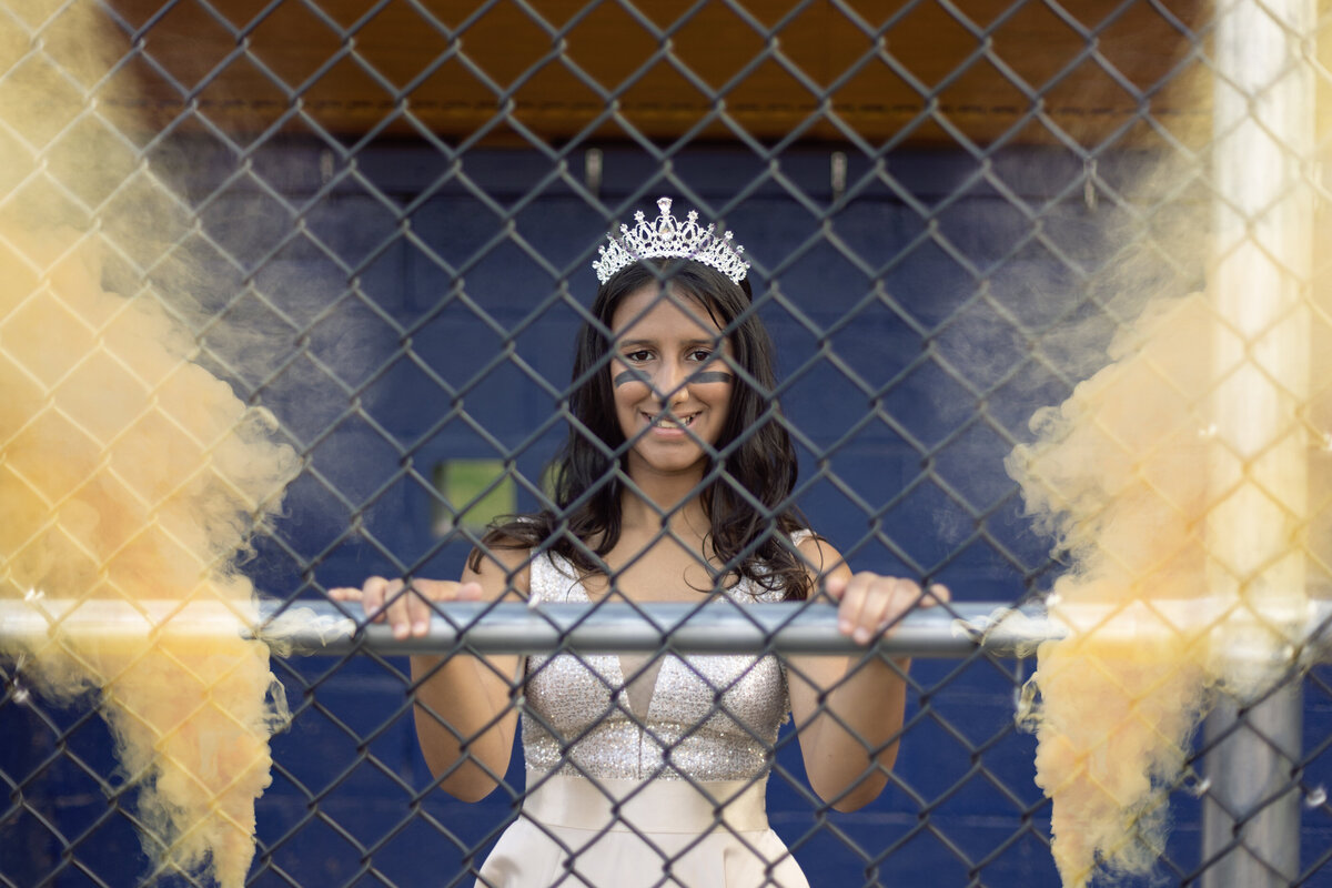 A high school girl in a dress and tiara stands behind a fence with powder cannons blowing on either side