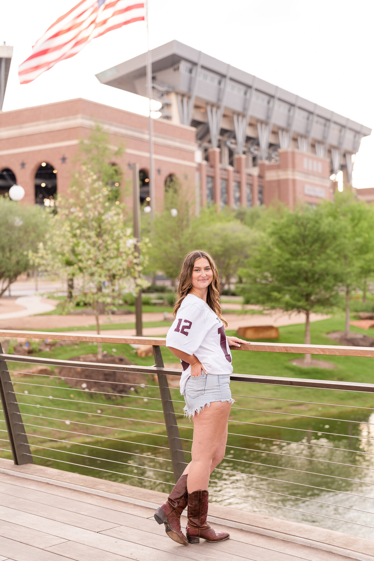 Texas A&M senior girl standing on bridge with hand on railing and other in back pocket while smiling over shoulder in Aggie Park with Kyle Field in background