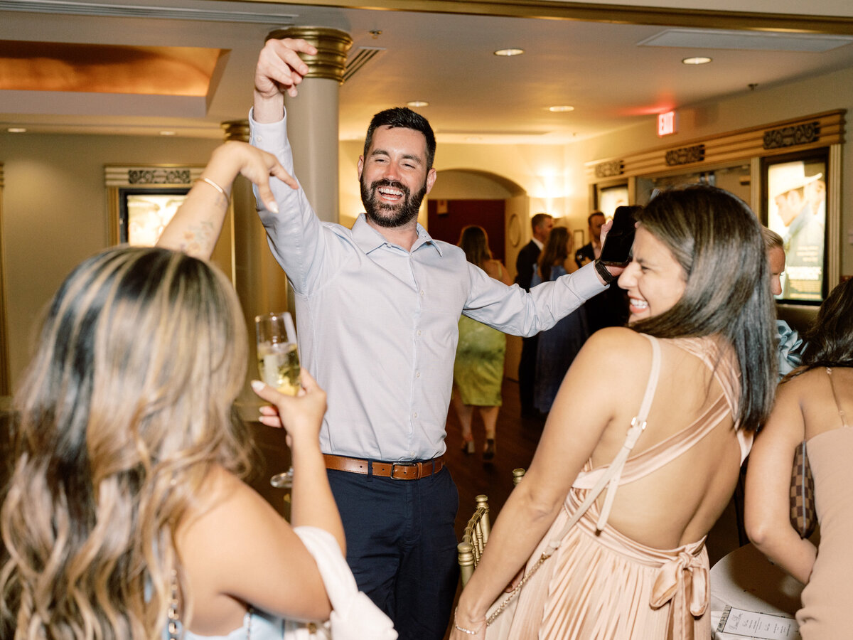 A man cheerfully raising his arms in celebration, surrounded by friends enjoying cocktails and the festive atmosphere of a vibrant social wedding.
