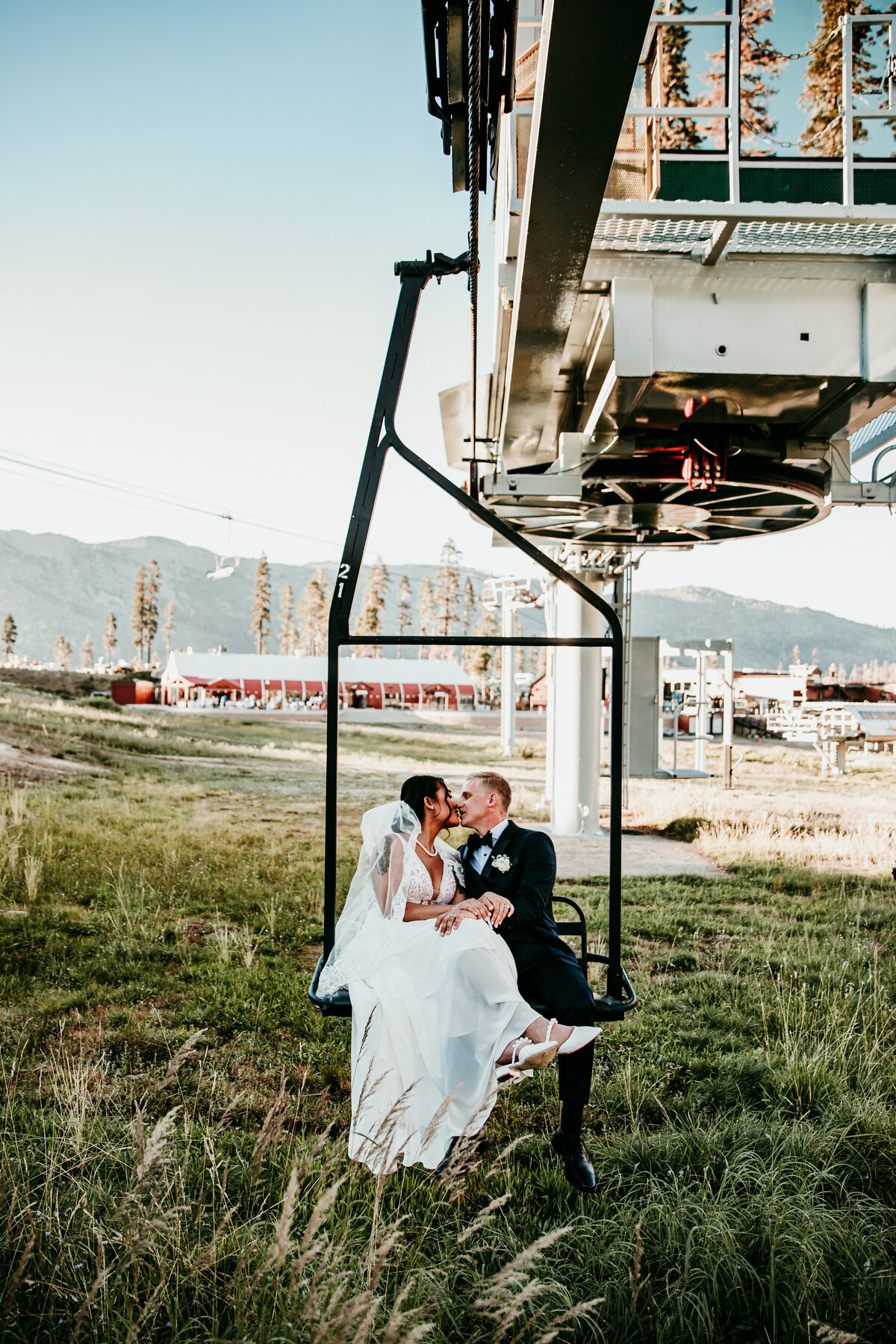 Bride and groom sharing a kiss while sitting on a ski lift at a mountain resort, with grassy fields and distant peaks in the background.