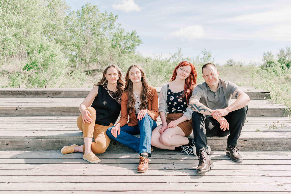 Mom, dad and daughters sit on a wooden walkway. 