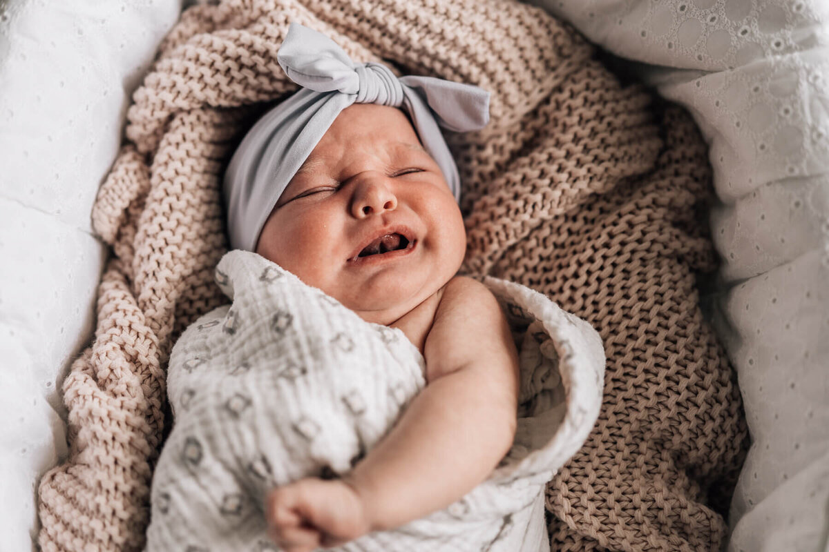A  newborn girl with a big bow headband begins to cry while lying on her back in a Moses blanket with a chunky knit textured blanket.