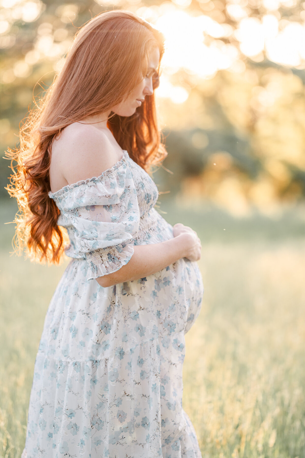 A pregnant woman, wearing w white dress with blue flowers, stands in a field of grass and holds her belly. The setting sun is glowing behind her.