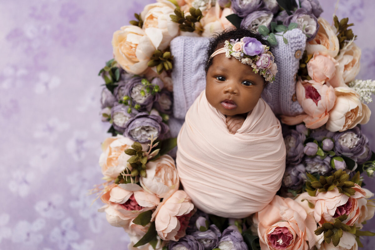 Baby girl wrapped in peach surrounded by purple florals during studio newborn session.