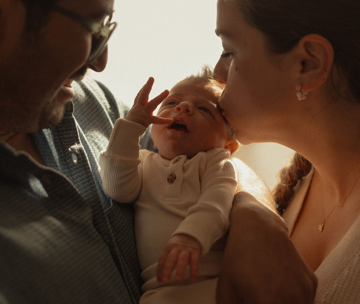 mother and father holding and kissing newborn baby in golden light