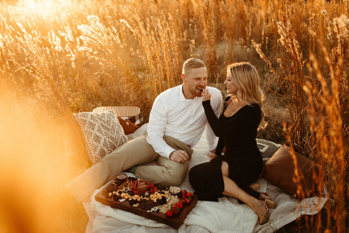 Couple sitting down on a picnic blanket in a field at sunset eating a charcuterie board during their engagement session