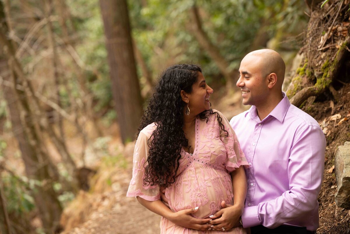 a couple stand in the woods, both wearing different shades of pink, as she holds her baby bump