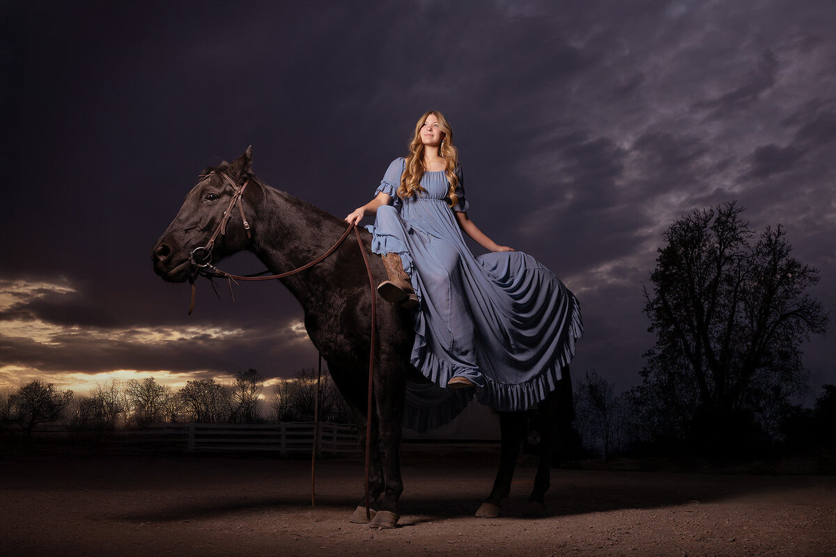 A senior girl in a flowing blue dress poses on her horse, captured by a Dallas senior photographer during a dramatic sunset.