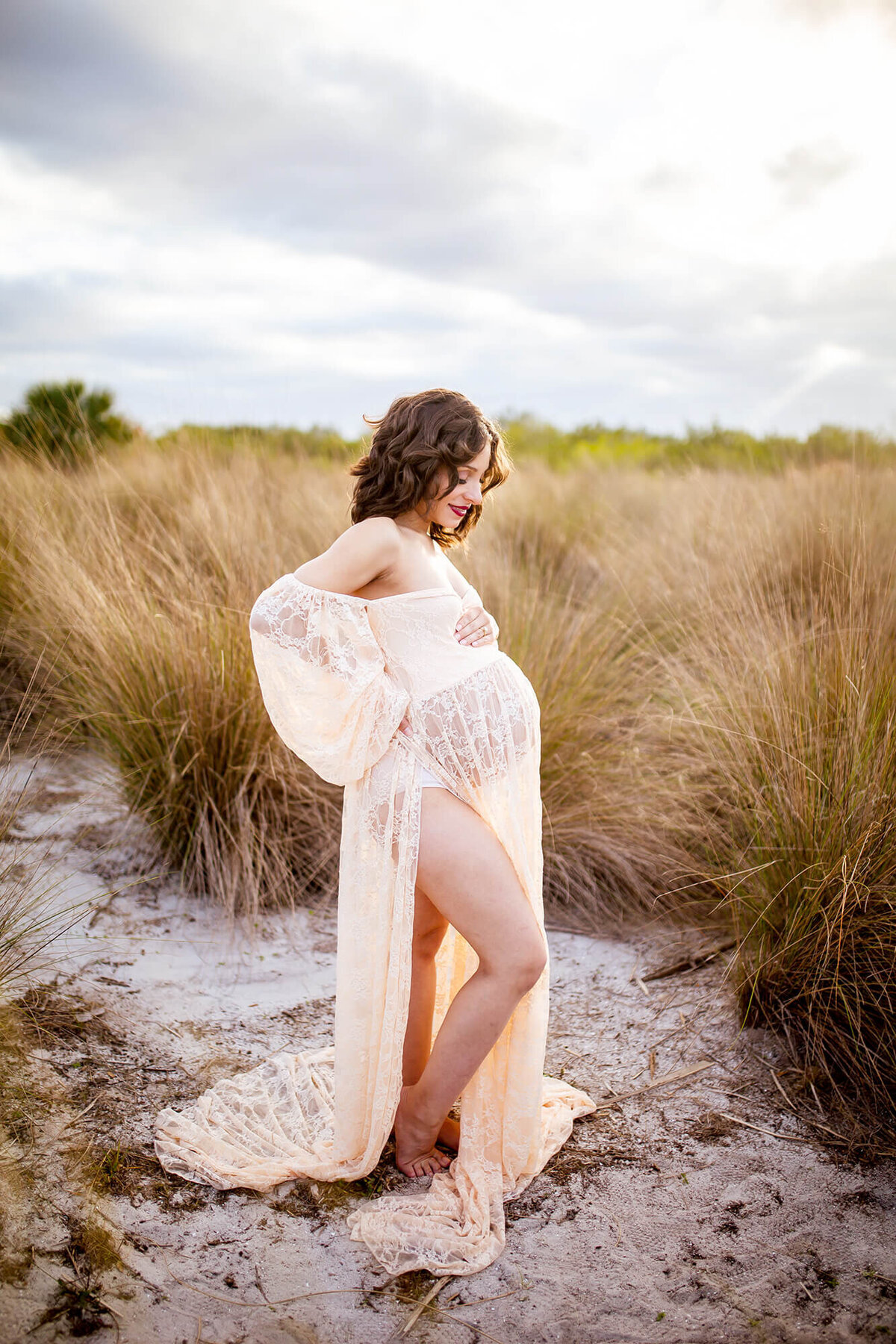 Pregnant dark haired woman standing in the middle of tall golden grass holding belly while looking down at belly. Cypress Point park 