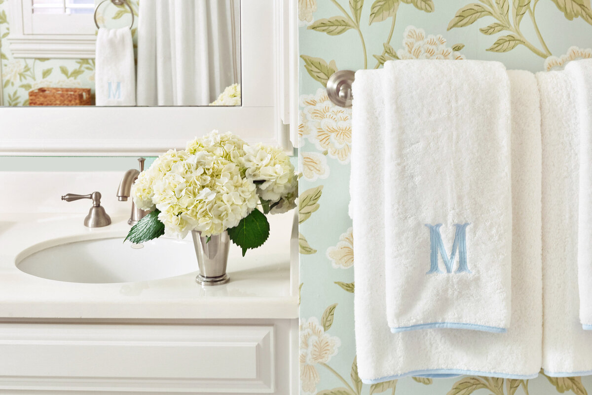 White bath towels with blue embroidery of initials with floral wallpaper and decorative floral arrangement on bathroom vanity