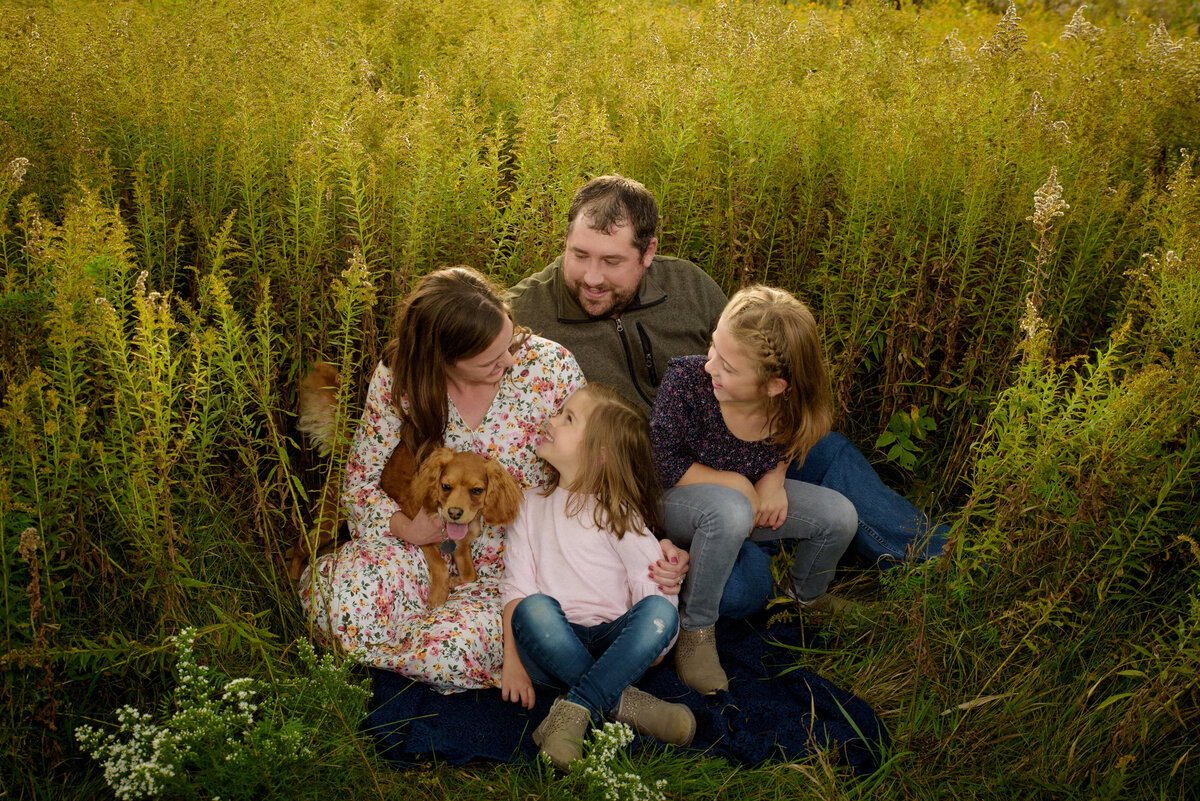 Portrait of a family with their little dog looking at each other and laughing in a long grassy field at Fonferek Glen County Park near Green Bay, Wisconsin.