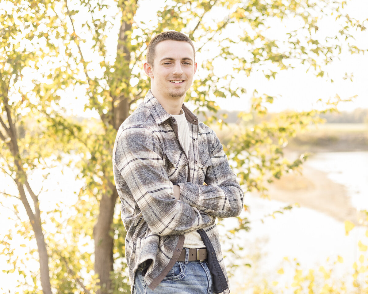high school senior male standing to the side in grass with trees behind him, arms crossed, wearing a flannel jacket, light tan shirt, blue jeans, and bel, smiling at the camera