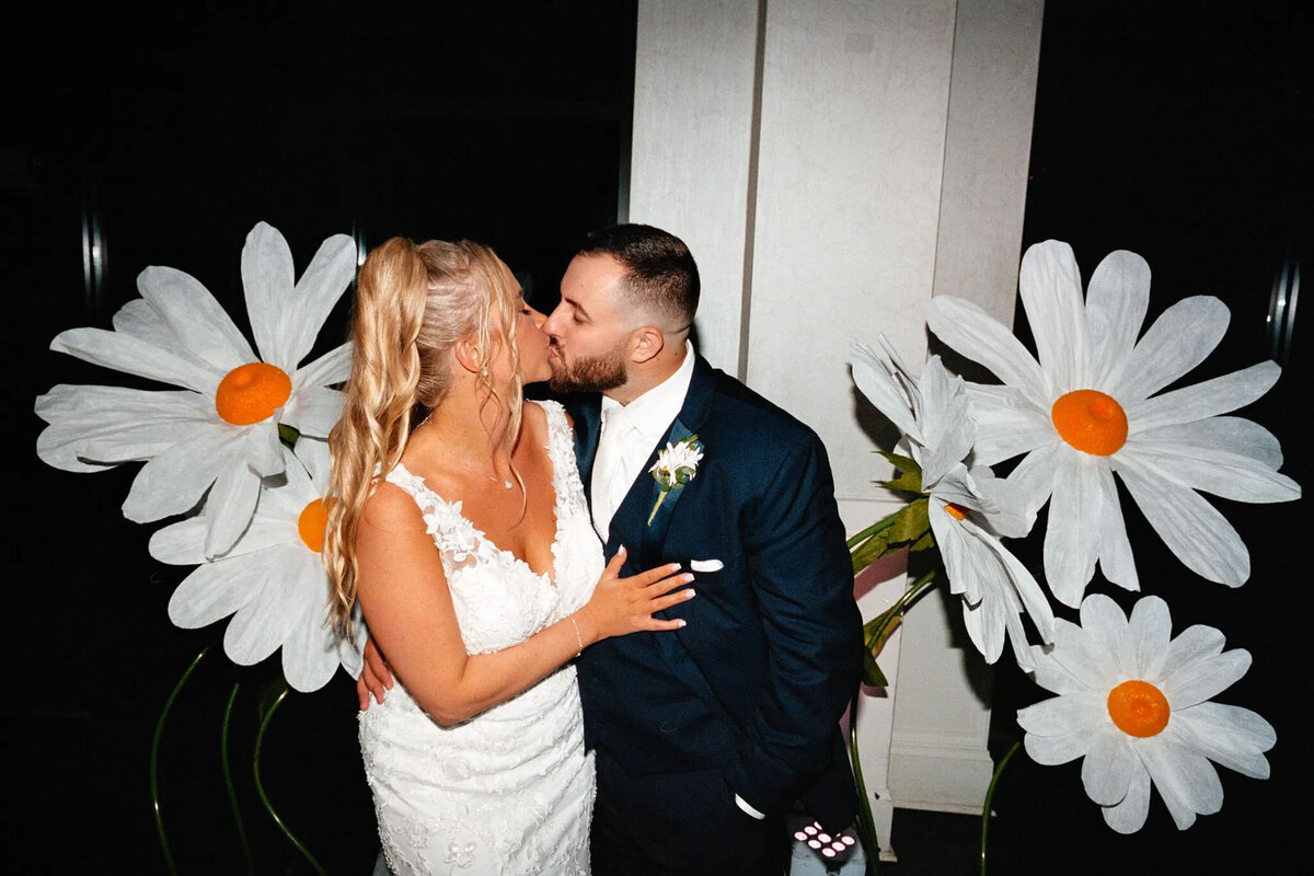 A bride and groom kiss in front of large, decorative white daisies with orange centers. Captured by a talented NJ wedding photographer, the bride stuns in a lace dress while the groom sports a dark suit with a boutonniere.