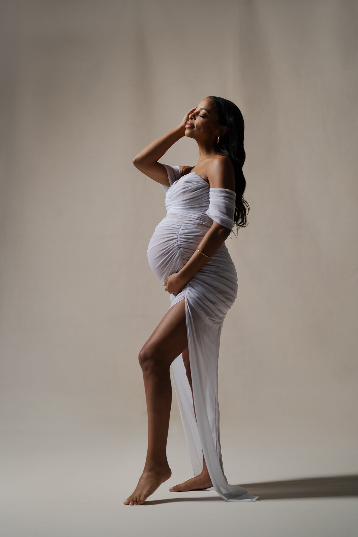 A full-body studio portrait of a mother-to-be wearing a flowing white dress, exuding elegance and strength in a professional photography session in NYC.