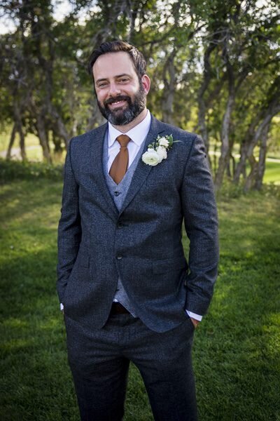 A groom stands with his hands in his pockets, smiling at the camera at The Oaks at Plum Creek in Denver, Colorado.