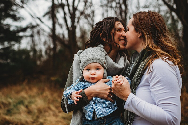 Family photographers Maryland captures LGBTQ+ family photos with mother kissing one another as they hold they toddler son for outdoor family pictures in the fall