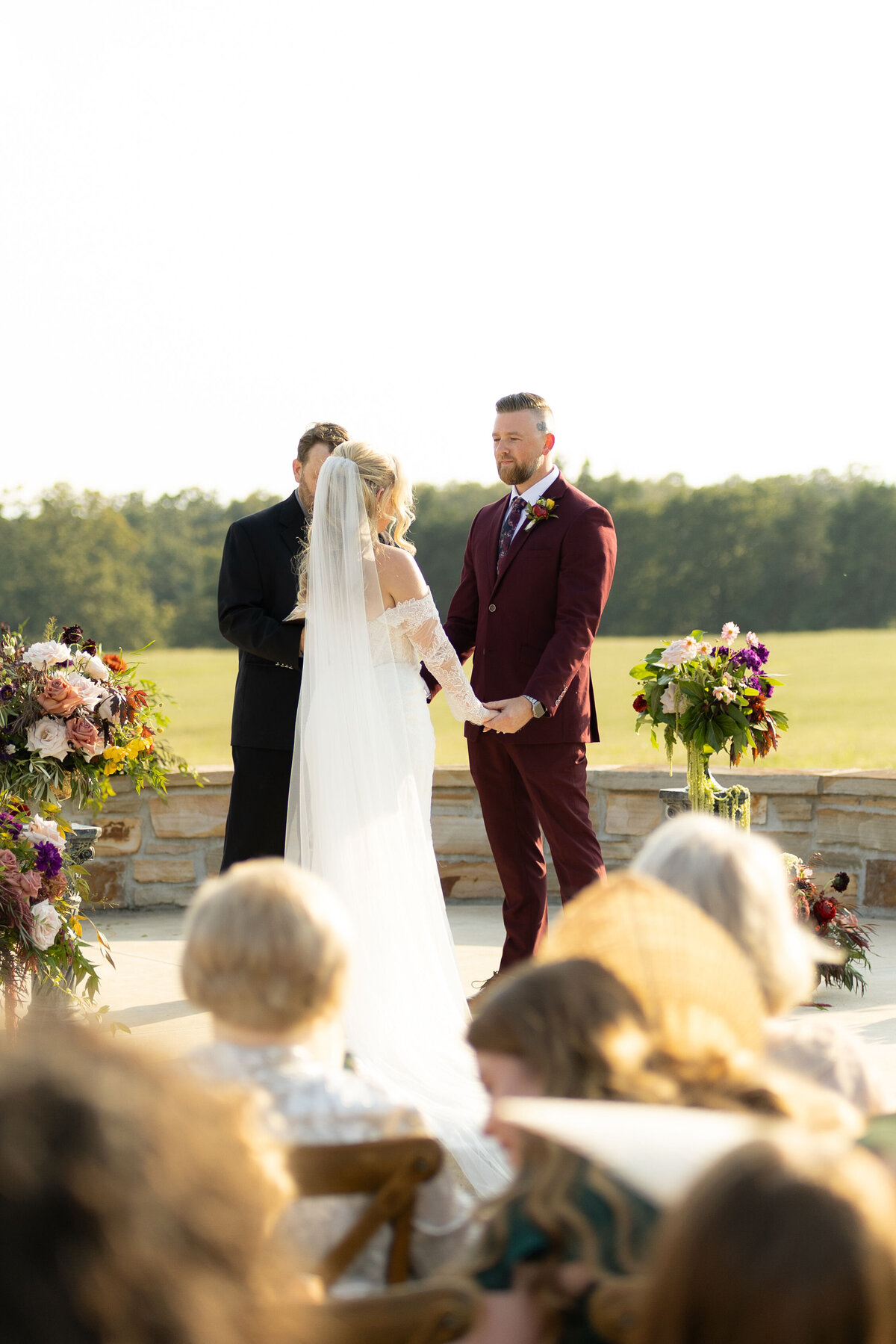 Bride and groom standing at an outdoor wedding ceremony surrounded by lush floral arrangements featuring mauve roses, burgundy blooms, and vibrant garden textures, designed by a wedding florist specializing in romantic, organic floral installations for elegant countryside weddings.