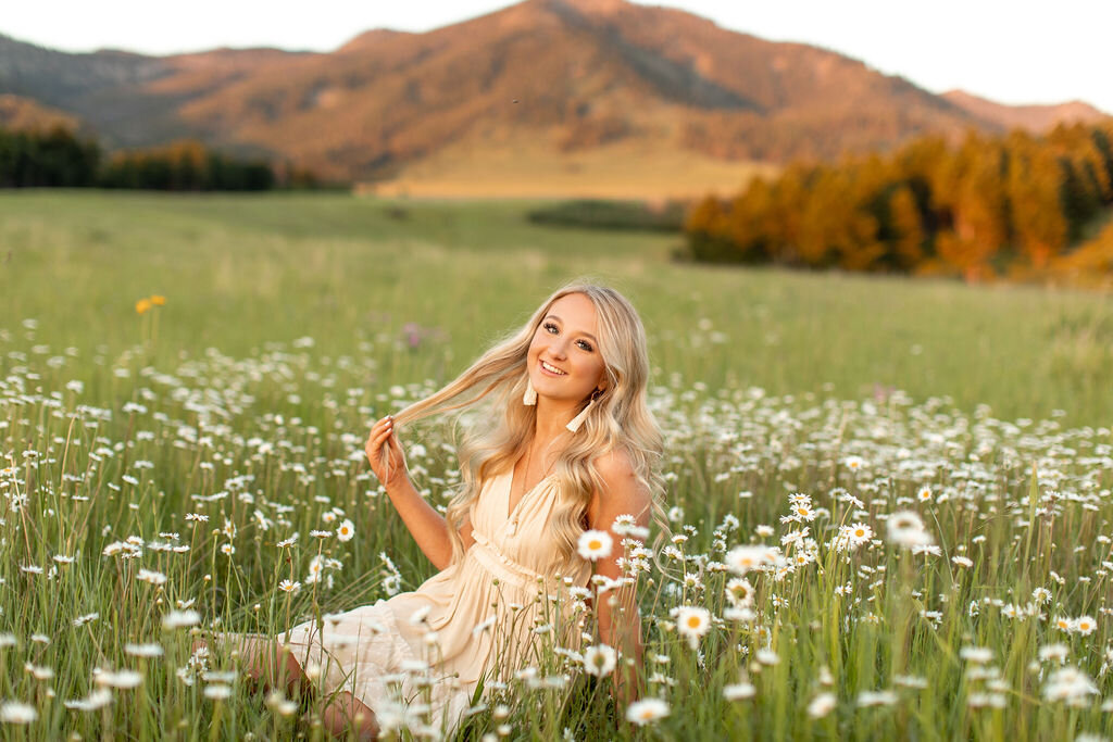 Young woman sits on rocks in a field with large trees as she rests her elbow on her knee.