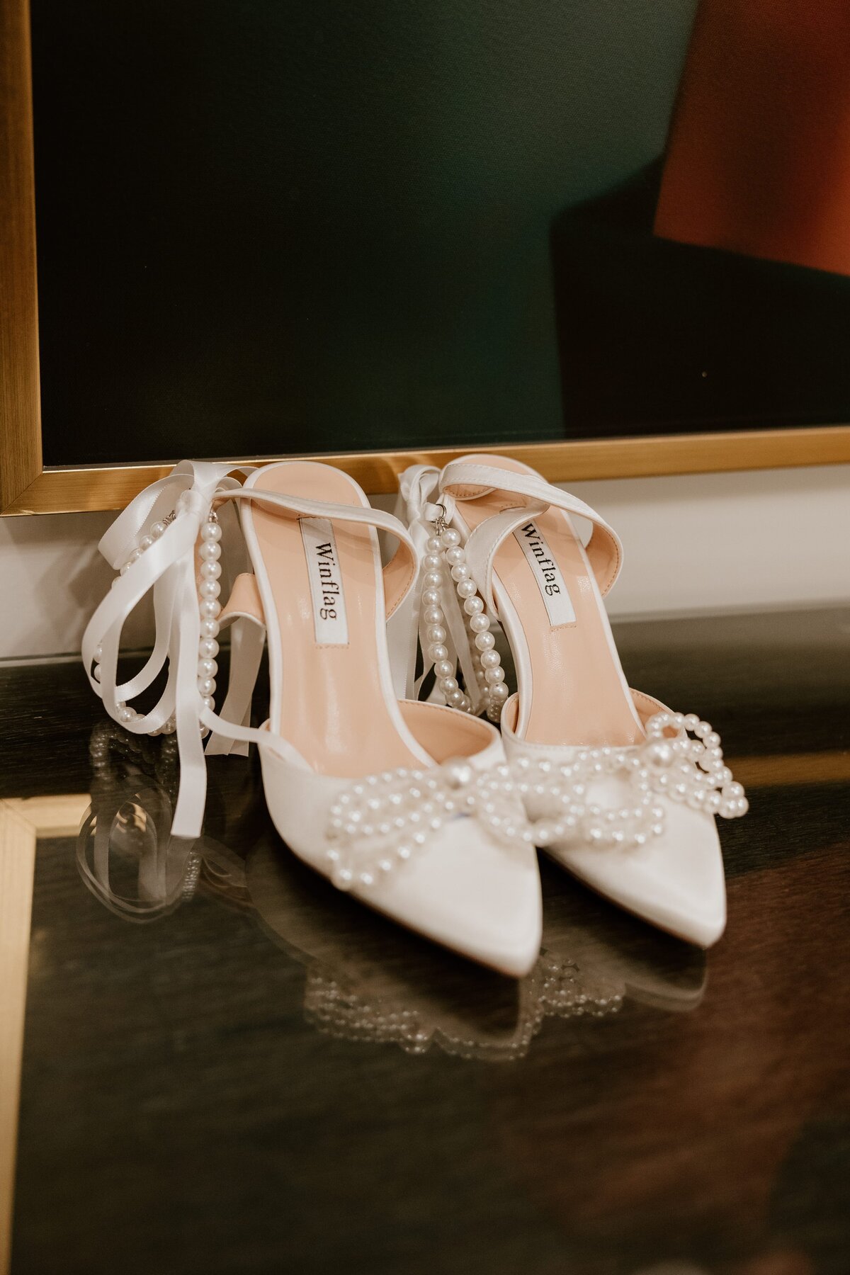 Elegant bridal shoes adorned with pearl bows and ribbon straps, displayed on a reflective glass surface at The Grandview in Poughkeepsie. A stylish detail shot from a Hudson Valley wedding.