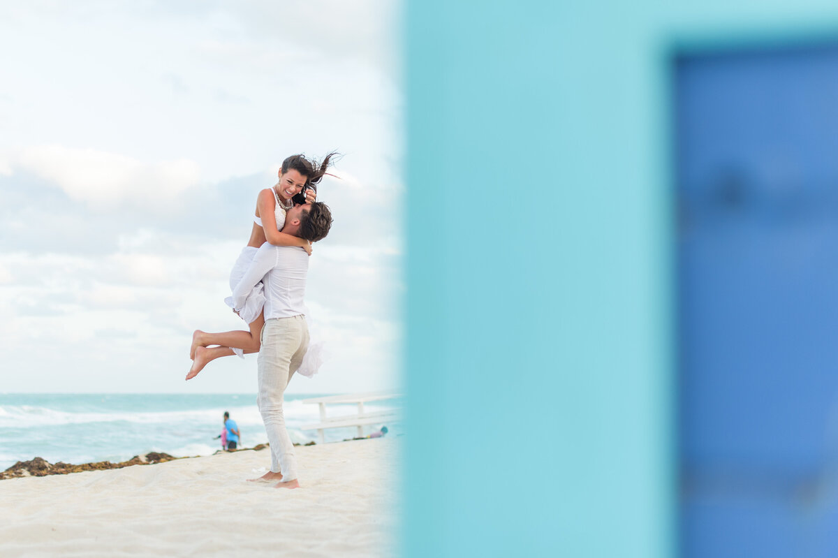 Engaged couple embracing on a Miami beach, with the man lifting the smiling woman against an ocean backdrop and bright sky, partially framed by a blue wall.