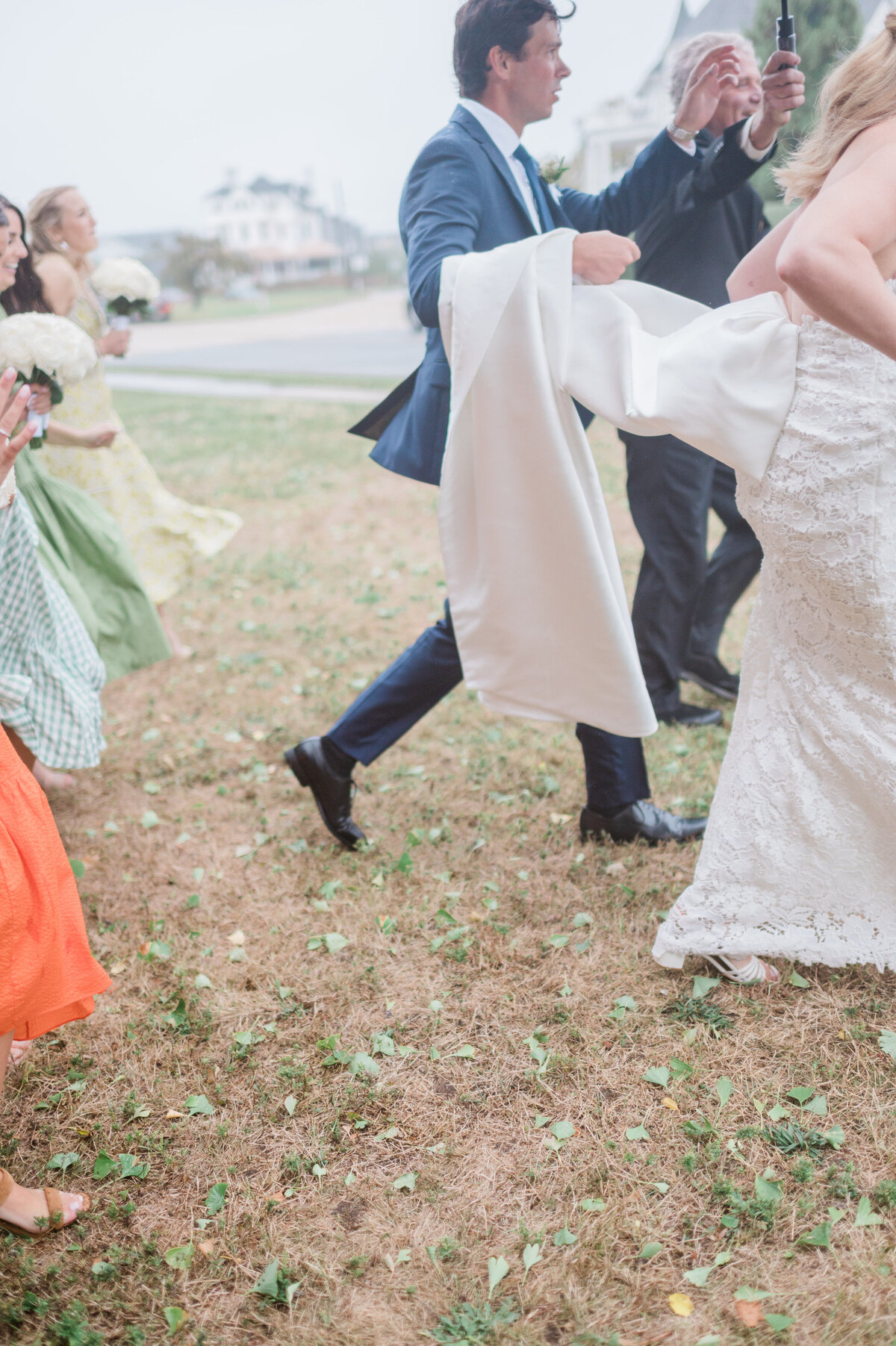 The groom, wearing a navy blue suit, walks briskly alongside the bride, who lifts the train of her lace wedding gown as they hurry to shelter.