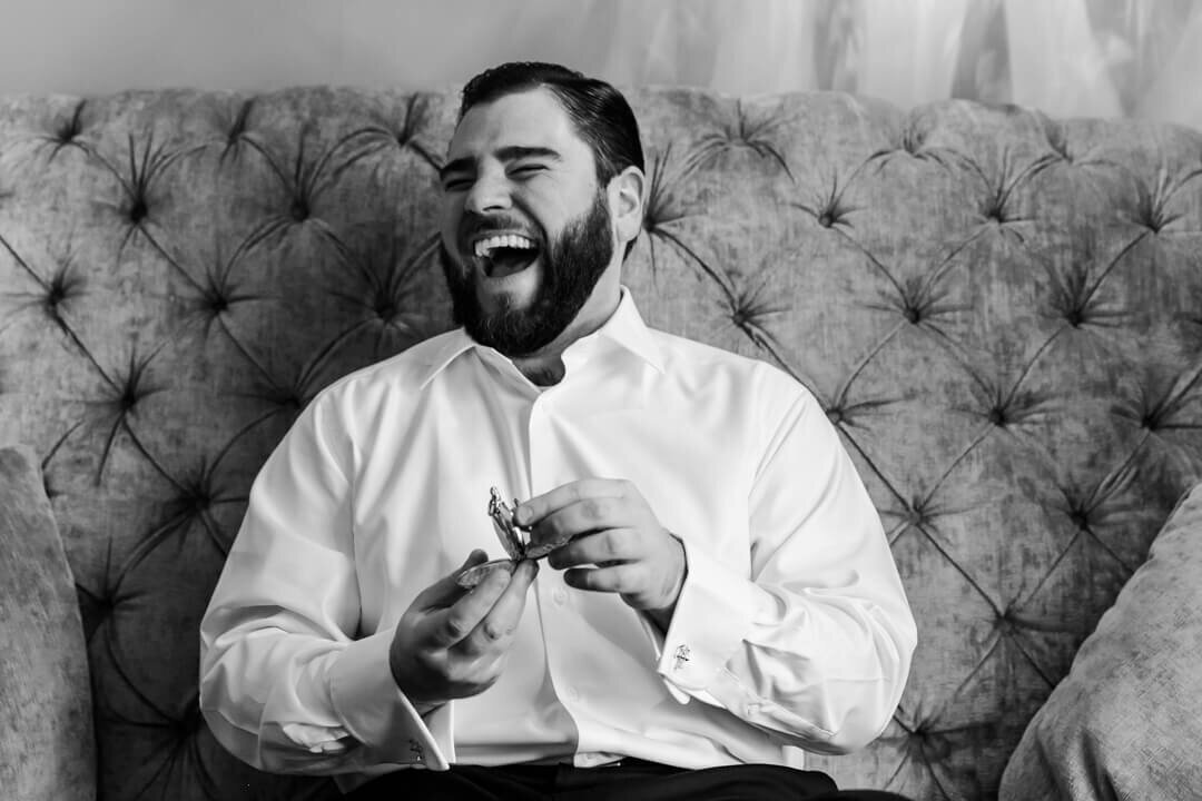 A groom sits in a large chair while getting ready and laughing in black and white