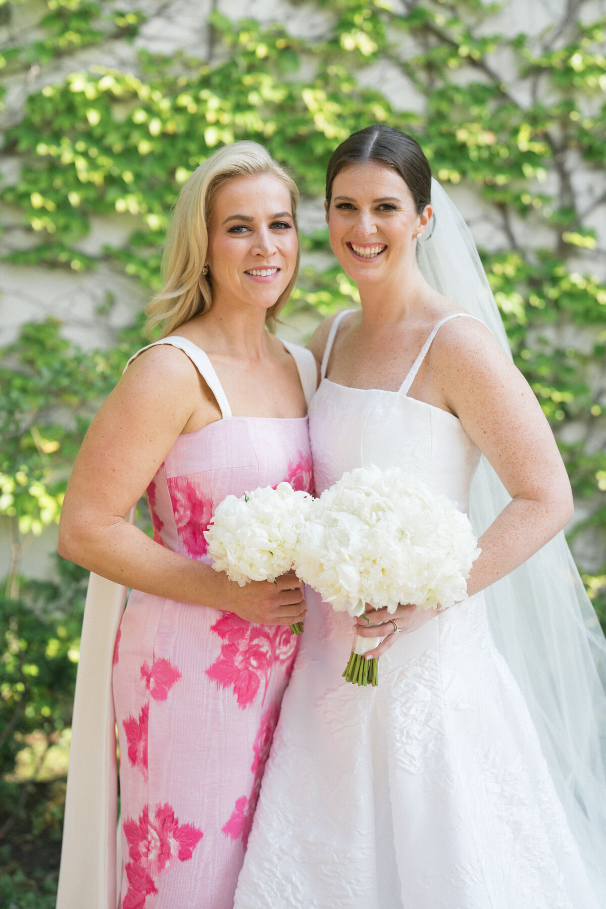Bride smiling alongside a bridesmaid in a pink dress, both holding white bouquets.